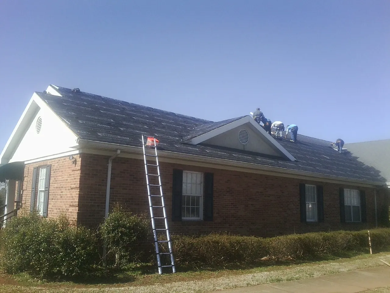 Crew tearing shingles off a brick ranch roof with a ladder set — Emergency Roof Repair work by our crew in Henrieville
