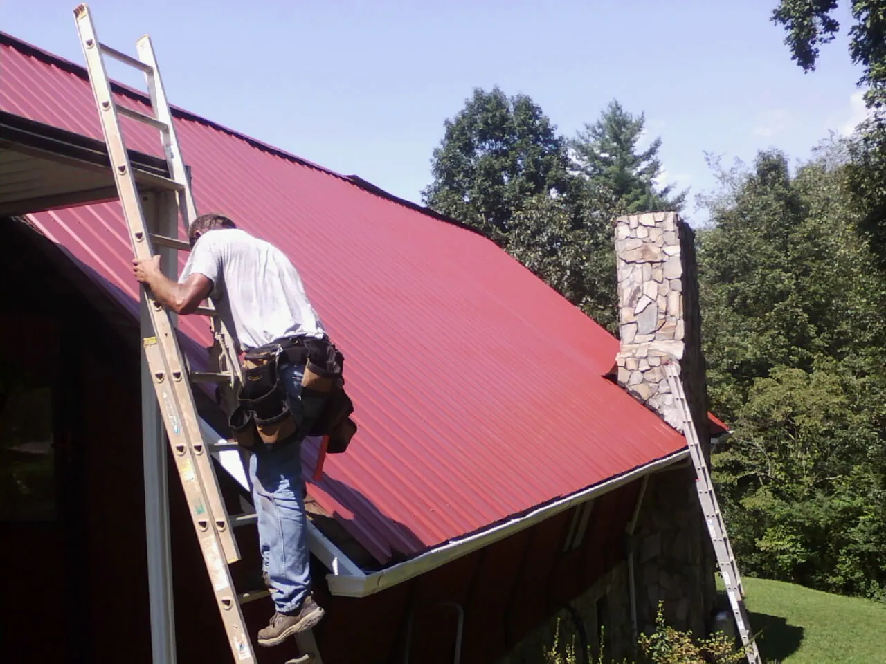 Roofer on a ladder beside a red metal roof and stone chimney — Soffit & Fascia Repair by Roofing Brothers in Henrieville